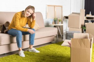 smiling young woman sitting on sofa while packing cardboard boxes during relocation