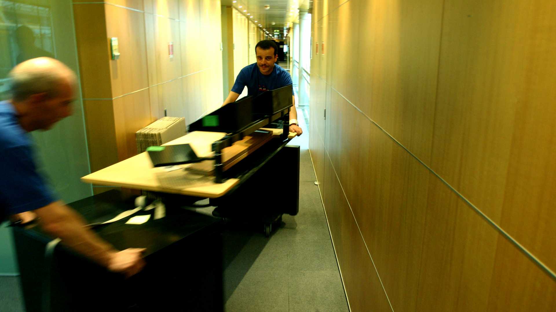 Deux hommes déplacent un meuble et des équipements dans un couloir d bureau.